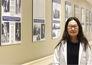 Photo de Minna Woo avec, en arrière-plan, des photos de l’exposition sur les 100 ans de la découverte de l’insuline. Photo prise dans le corridor de la Banting and Best Diabetes Clinic à l’Hôpital général de Toronto, où la première dose d’insuline a été administrée à Leonard Thompson il y a 100 ans, un an seulement après la découverte de l’insuline. 