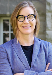 Greta Bauer, Ph. D., une personne à la peau claire, aux cheveux mi-longs et châtains avec des reflets blonds, est à l'extérieur par beau temps. Elle porte des lunettes rondes, noires et violettes, ainsi qu’un blazer violet par-dessus un chandail noir. Elle regarde vers la caméra et sourit avec les bras croisés.